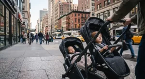 Parent pushing a double stroller for one infant and one toddler along a busy downtown city sidewalk.
