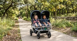 Side-by-side double stroller with equal-size seats featuring all-terrain wheels on a park trail sidewalk.