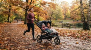 A parent pushing an all-terrain twin jogging stroller for trail runs through a park covered in autumn leaves.