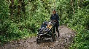 An all-terrain twin jogging stroller for trail runs tackling a muddy section of a national park hiking trail.