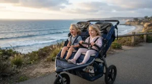 Two happy toddlers smiling inside a jogging double stroller for marathon training parents on a sunny coastal boardwalk
