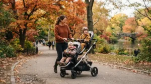A sit-and-stand stroller for toddler and preschooler being pushed along a sunny park pathway.