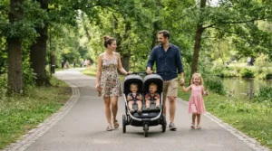 A family enjoying an afternoon stroll on a paved urban park path using a twin stroller for narrow city sidewalks.